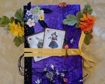 Puede incluir: Un diario de terciopelo morado decorado con naipes, abalorios y flores. El diario presenta imágenes de Alicia en el país de las maravillas, con una cinta amarilla y abalorios decorativos. La portada está adornada con flores artificiales y una cinta negra.
