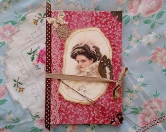 Puede incluir: Un diario hecho a mano con una cubierta de patrón floral rosa, adornado con encaje, un retrato vintage y un acento de mariposa. El diario está atado con una cinta y adornado con elementos decorativos.