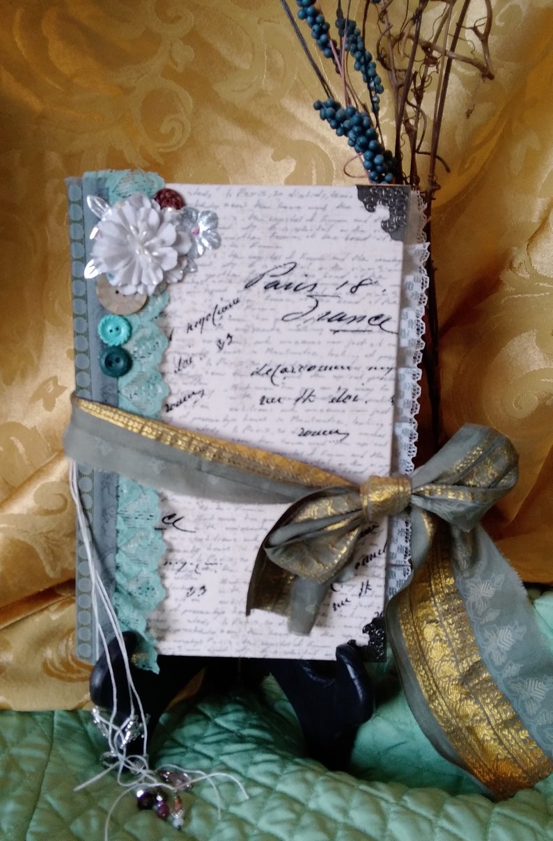 Puede incluir: Un diario decorativo con est&eacute;tica vintage. La portada presenta escritura manuscrita, ribete de encaje y un lazo de cinta gris y dorada. Los adornos incluyen botones y una flor blanca. El texto "Paris 18 France" es visible.