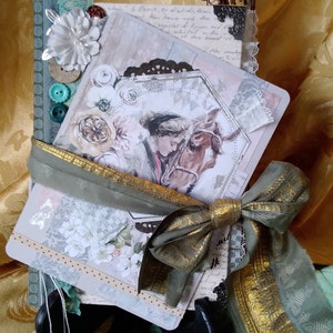 Puede incluir: Un diario decorativo con una imagen de caballo y jinete, adornado con una cinta verde salvia con detalles dorados. El diario presenta adornos florales, botones y encaje. Una ramita de bayas y ramas secas est&aacute;n en el fondo.