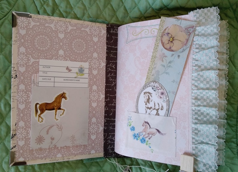 Puede incluir: Diario abierto con una pegatina de caballo y un marcap&aacute;ginas. El diario tiene un patr&oacute;n floral rosa y un ribete de encaje. El marcap&aacute;ginas presenta una ilustraci&oacute;n de caballo y una flor. El diario tiene una etiqueta con campos para autor, t&iacute;tulo, fecha de vencimiento y nombre del prestatario.
