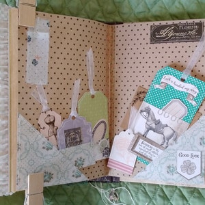 Puede incluir: Scrapbook abierto con p&aacute;ginas decorativas, bolsillos y etiquetas. Las p&aacute;ginas tienen un patr&oacute;n de lunares. Las etiquetas con cintas son de varios colores y formas. Una etiqueta dice "Luck is headed my way". El &aacute;lbum de recortes se mantiene abierto con pinzas de madera.