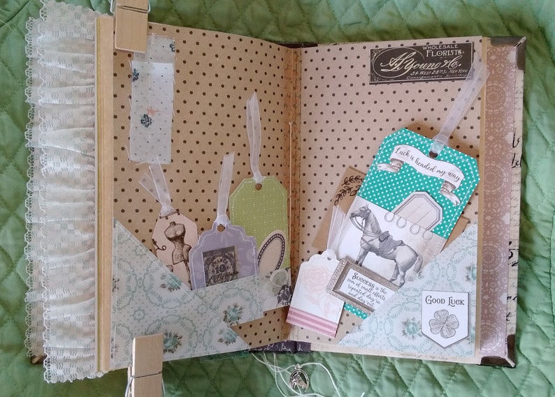 Puede incluir: Scrapbook abierto con p&aacute;ginas decorativas, bolsillos y etiquetas. Las p&aacute;ginas tienen un patr&oacute;n de lunares. Las etiquetas con cintas son de varios colores y formas. Una etiqueta dice "Luck is headed my way". El &aacute;lbum de recortes se mantiene abierto con pinzas de madera.