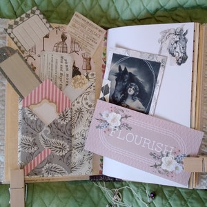 Puede incluir: Un diario hecho a mano con papel decorativo, ribete de encaje y ef&iacute;meras vintage. El diario presenta una tarjeta rosa con la palabra "FLOURISH" y una imagen en blanco y negro de una mujer y un caballo. El diario se cierra con una pinza de madera.