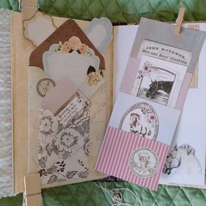 Puede incluir: Diario abierto con p&aacute;ginas decorativas, bolsillos y adornos. Presenta dise&ntilde;os florales y de estilo vintage, incluyendo un bolsillo con una ilustraci&oacute;n de caballo y el texto "JOHN MOTHNER, Men and Boys' Clothing." Incluye ribete de encaje y etiquetas de papel.