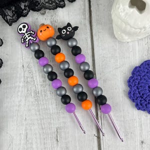 May include: Three Halloween-themed crochet hooks with purple, black and orange beads. The hooks feature a skeleton, a pumpkin and a bat.