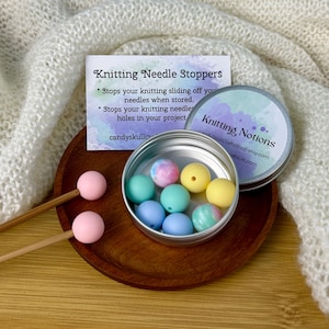 May include: A wooden bowl contains colourful knitting needle stoppers in a small tin. The tin lid reads "Knitting Notions." Two knitting needles with pink stoppers are on the left. A card reads "Knitting Needle Stoppers."