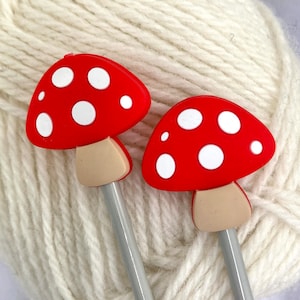 May include: Two red and white mushroom-shaped knitting needles with silver metal shafts. The needles are resting on a ball of white yarn.