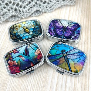 May include: Four rectangular metal pill boxes with decorative tops. The designs include a bee, a butterfly, a hummingbird, and a dragonfly. Each box has a silver frame and a hinged lid. The artwork features vibrant colours and intricate details.