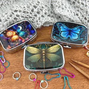 May include: Three decorative metal pill boxes with butterfly and dragonfly designs. The boxes feature vibrant colours and intricate details. Knitting accessories, including stitch markers and needles, are arranged around the boxes on a wooden surface.