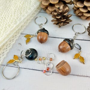 May include: Three wooden acorn-shaped stitch markers with metal key rings and charms. The charms are small golden leaves. One acorn is open, revealing a set of stitch markers inside.
