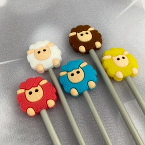 May include: Five knitting needles with sheep-shaped tops in different colours: red, white, brown, blue, and yellow. The needles are silver and have a decorative pattern.
