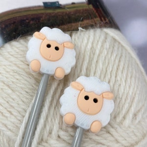 May include: Two grey knitting needles with white sheep-shaped tops. The sheep have black eyes and pink noses.