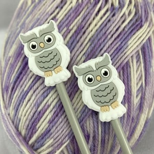 May include: Two grey and white owl-shaped knitting needle tips on grey knitting needles. The needles are resting on a ball of purple and white yarn.