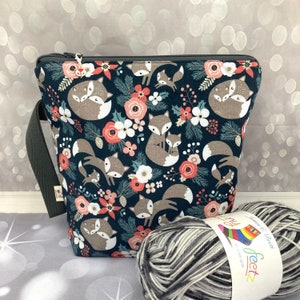 May include: A navy blue fabric bag featuring a fox and floral pattern. The bag has a grey strap and zip closure. Two balls of yarn are in front of the bag. The yarn is black, white, and grey.