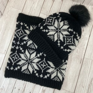Puede incluir: Conjunto de gorro y bufanda de punto negro y blanco con un patrón de copo de nieve. El gorro tiene un pompón de piel sintética negra.