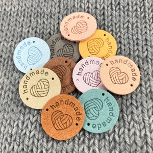 May include: Assortment of round, leather labels with the text "handmade" and a heart-shaped yarn ball design. The labels are in various colours, including light blue, brown, beige, pink, and green.
