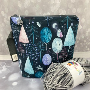 May include: A black zippered pouch with a woodland scene featuring trees, owls, and stars. The pouch is made of fabric and has a silver charm attached to the zipper. The pouch is sitting on a grey surface next to a ball of grey yarn with a white label that says "Funny" and "Summer Love".
