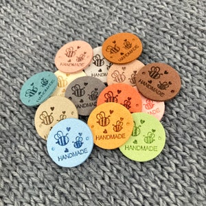 May include: A selection of small, circular tags in pastel shades. Each tag displays a bee design and the word "HANDMADE". The tags are made of a suede-like material and are scattered on a grey, textured background.
