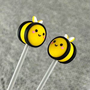 May include: Two yellow and black bee-shaped lollipops with white wings and pink cheeks on clear sticks.