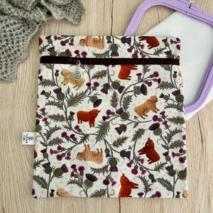 May include: A rectangular fabric pouch featuring a repeating pattern of brown Highland cattle and purple thistle flowers on a cream background. The pouch has a dark brown zip closure with a small circular metal charm attached to the zip pull.