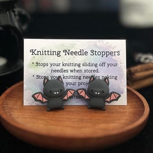 May include: Two black bat-shaped knitting needle stoppers with pink wings are shown on a wooden plate. A card behind them reads "Knitting Needle Stoppers" and describes their function. The background is dark.