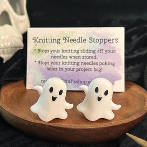 May include: Two white ghost-shaped knitting needle stoppers with black eyes and mouths sit on a wooden plate. A card behind them reads "Knitting Needle Stoppers". The background includes a skull and black fabric.