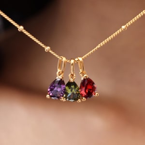 May include: A gold necklace featuring three teardrop-shaped pendants with purple, green, and red gemstones. The necklace has a delicate chain and is worn around the neck. The gemstones are vibrant and eye-catching.