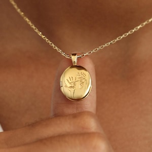 May include: A gold chain necklace with a gold oval locket pendant featuring an engraved handprint design.