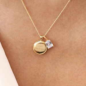 May include: A gold-colored necklace with two pendants. One pendant is a round disc with the engraved text "Love you forever". The other pendant is a small, clear, rectangular gemstone. The necklace is worn against a white background.