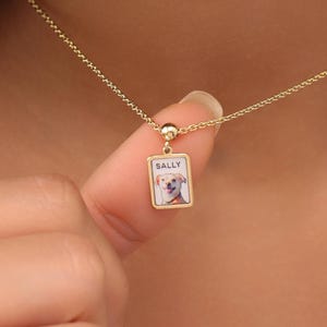 May include: A gold-colored necklace with a small, square pendant. The pendant features a picture of a dog and the name "SALLY" in white text. The necklace is delicate and appears to be worn around the neck.