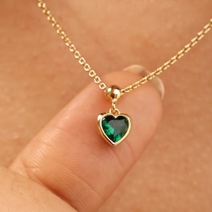 May include: A gold-colored necklace with a delicate chain and a heart-shaped pendant. The pendant features a vibrant green gemstone set in a gold bezel. The necklace is displayed against a skin-toned background.