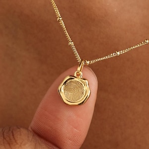 May include: A gold-colored necklace with a fingerprint pendant. The pendant is round with a detailed fingerprint design. The necklace is on a person's neck and finger.