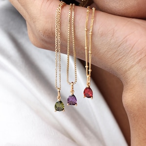 May include: Three gold necklaces with teardrop-shaped pendants in green, purple, and red gemstones. The necklaces are displayed against a white background. The chains have different designs, including a box chain and a beaded chain.