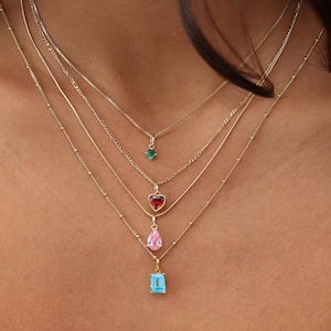 May include: A collection of gold necklaces with gemstone pendants. The pendants include a green round stone, a red heart-shaped stone, a pink teardrop, and a blue rectangular stone. The necklaces are layered and vary in chain style.