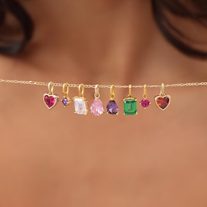May include: A delicate gold chain necklace featuring seven colorful gemstone charms. The charms include heart, emerald cut, and pear shapes in various colors like pink, purple, and green. The necklace is displayed against a blurred background.