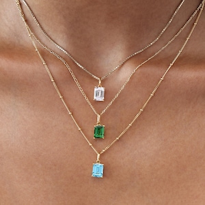 May include: Three gold necklaces with pendant gemstones. The top necklace has a clear gemstone, the middle one has a green gemstone, and the bottom one has a blue gemstone. The necklaces are layered and worn close to the neck.