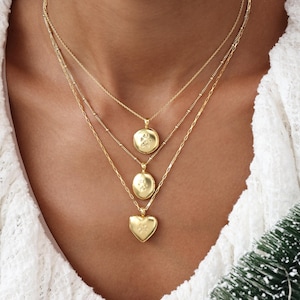 May include: Three gold necklaces with pendant charms. The top two pendants are round, and the bottom pendant is heart-shaped. Each pendant has a small engraved design. The necklaces are layered and vary in chain style.