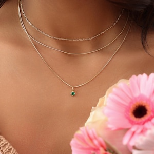 May include: A layered gold necklace set. The top two necklaces are delicate chains, and the bottom necklace features a small, green gemstone pendant. The image is set against a soft, neutral background, with a bouquet of pink flowers in the foreground.