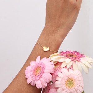 May include: A delicate gold bracelet featuring a heart-shaped charm with a floral design. The bracelet is worn on a wrist, with a bouquet of pink and white gerbera daisies in the foreground. The bracelet's chain is thin and elegant.