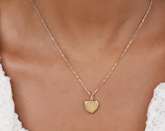 May include: A gold-colored necklace with a heart-shaped pendant. The pendant has a textured surface and hangs from a delicate chain. The necklace is worn against a white background.