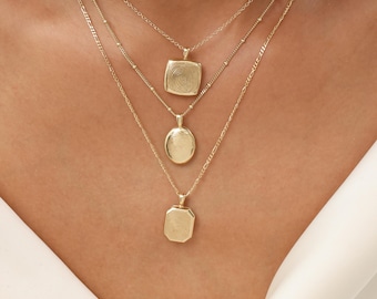 May include: Three layered gold necklaces featuring pendants with engraved fingerprint patterns. The top pendant is square, the middle is oval, and the bottom is rectangular. Each pendant hangs from a delicate gold chain of varying styles.