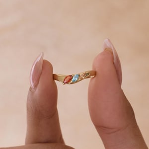 May include: A gold ring featuring a curved band set with three marquise-cut gemstones arranged in a row. The stones consist of a deep orange-red gem, a bright light blue gem, and a pale translucent gem, all securely mounted in a polished gold setting.