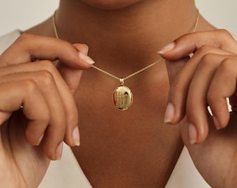 May include: A gold-colored oval locket pendant necklace. The locket has an engraved design and hangs from a delicate chain. The necklace is held up by a person's hands.