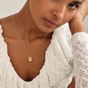 May include: A gold necklace featuring a small, oval pendant suspended from a fine chain. The necklace is shown against a white, textured top. The pendant is a warm gold colour.