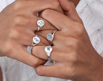 May include: Four polished silver signet rings featuring distinct engravings. One ring displays a floral bouquet, another a single flower, one a detailed fingerprint pattern, and the final ring features a stylized letter H in a cursive script font.