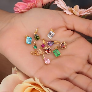 May include: A collection of gemstone pendants in various shapes and colors, including blue, green, purple, and red. Each pendant has a gold-colored bail. The pendants are displayed on a hand, with flowers in the background.