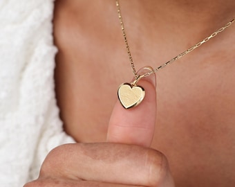 Puede incluir: Un collar con colgante de corazón dorado. El colgante en forma de corazón presenta un diseño de huella dactilar. El collar tiene una cadena fina. La imagen es un primer plano, que destaca los detalles del colgante y la textura de la cadena.