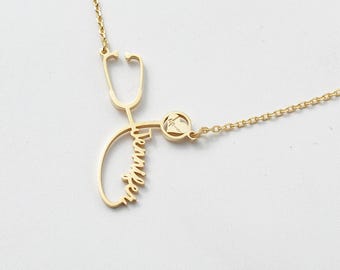 May include: A gold-colored necklace featuring a stethoscope design with the name "Jennifer" in cursive script. The necklace includes a small, round charm with a gemstone and a delicate chain.