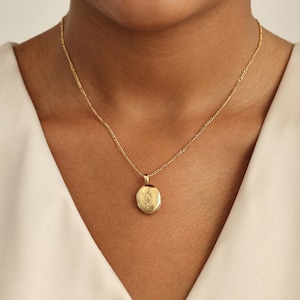 May include: A gold necklace with a fine chain and an oval locket pendant. The locket has an engraved floral design. The necklace is worn against a cream-coloured top.